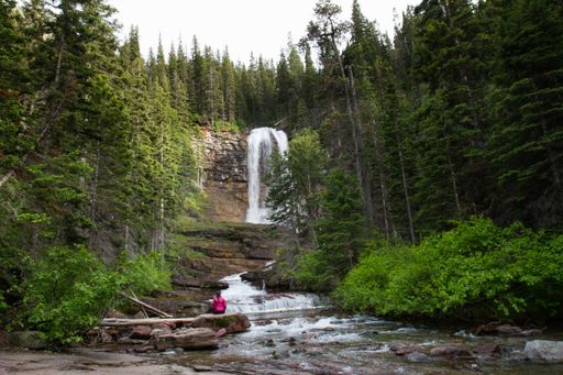 A female sitting on a rock looking at the waterfall surrounded by trees shot from behind