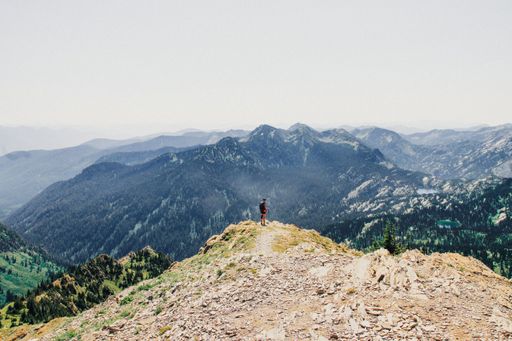 A beautiful shot of a person standing on edge of the cliff with forested mountains in the background