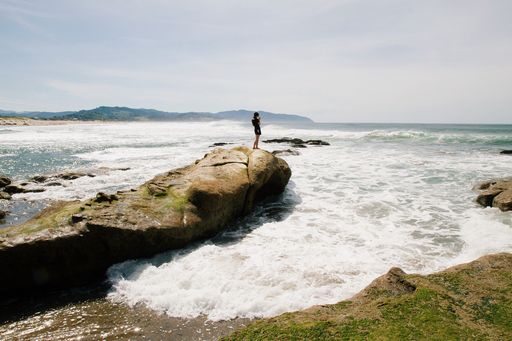 A beautiful shot of a female standing on a rock near the sea with mountains and a clear sky in the background