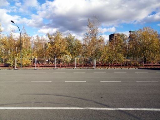 Empty parking lot with white lines, blue sky, clouds, trees, and building in the background
