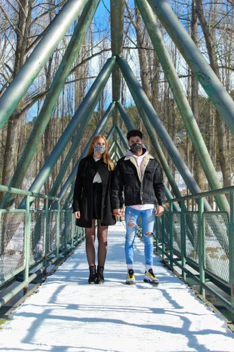 A vertical shot of a young interracial couple of tourists with masks walking on a snowy bridge