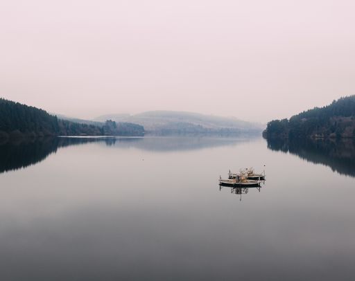 A high angle shot of the reflection of boats on a foggy lake surrounded by tree-covered hills