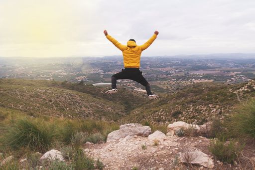 The man in a yellow down jacket jumping on the edge of the cliff against the sky.