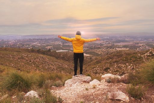 The man in a yellow down jacket and black pants standing on the edge of the cliff with stretched hands.