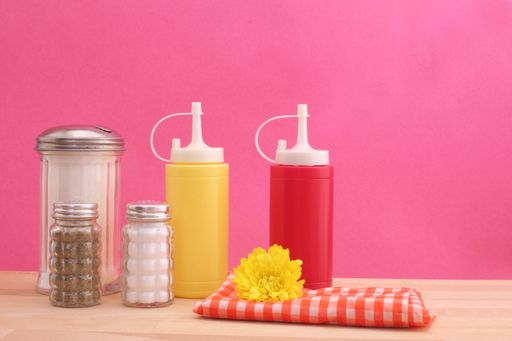 A closeup of ketchup, mustard, sugar, pepper, napkin with a yellow flower against the pink background.