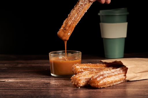 A close up shot of dipping churro in dulce de leche caramel. Wooden table, mug of coffee in backgroud