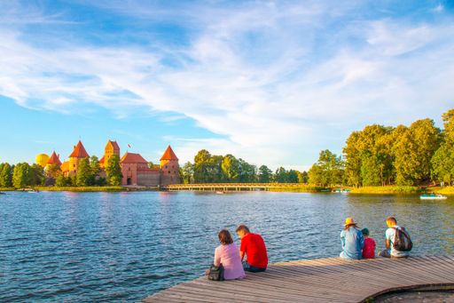 A view of tourists sitting on the lakeshore with trees and Trakai Island Castle on the background in Lithuania