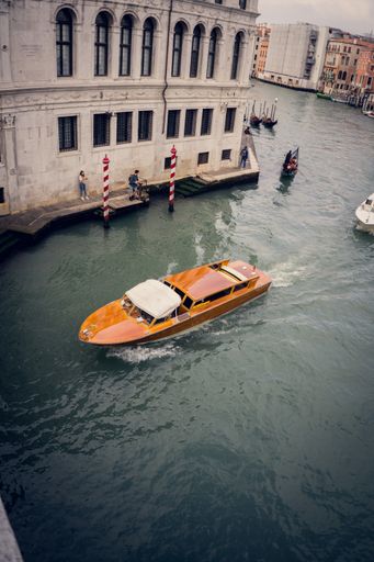 A vertical picture of an orange boat sailing on the grand channel next to the building in Venice, Italy