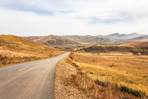 A road between golden fields surrounded by mountains under a cloudy sky