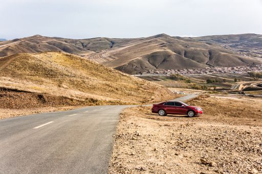 A car parked by a road between golden fields surrounded by mountains under a cloudy sky