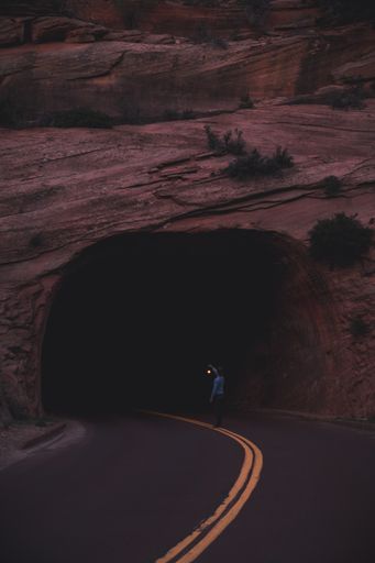 A vertical shot of a person holding a lantern standing in the middle of a road by a tunnel