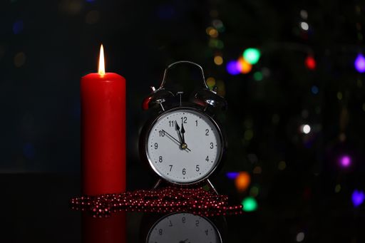 A close-up shot of a burning candle and an alarm clock with a lit Christmas tree in the background