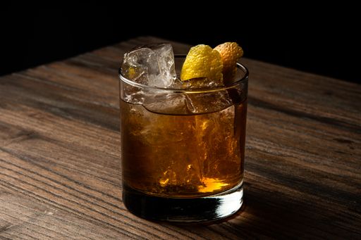 A closeup of a glass of old fashioned cocktail with ice on a wooden table with a blurry background