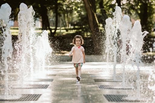 A shot of the little Caucasian girl playing in the park and running between jets of water in summer