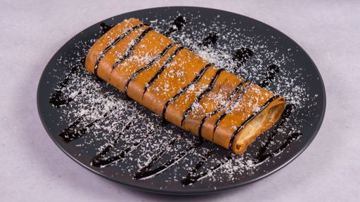A close-up shot of sweet crepe topped with chocolate sauce and coconut flakes in a gray plate on a white table