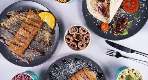 A top view of sweet crepes and tortilla in gray plates and ice cream in different flavors with fork and knife on a white table