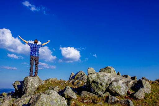 A back view of a man standing on the rock with open arms against a blue cloudy sky on a sunny day