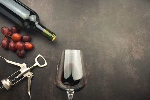 A top view of a red wine glass and bottle on a stone background with a corkscrew and grapes