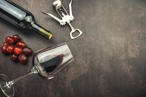 A top view of a red wine glass and bottle on a stone background with a corkscrew and grapes