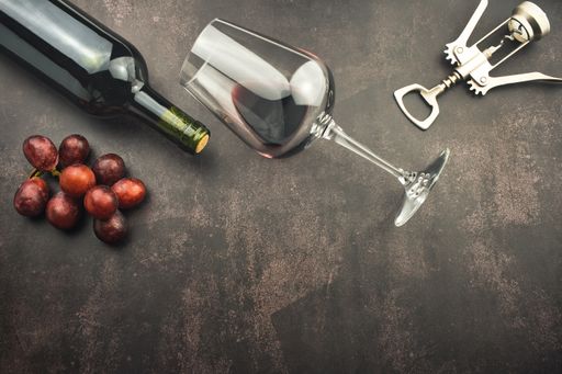 A top view of a red wine glass and bottle on a stone background with a corkscrew and grapes