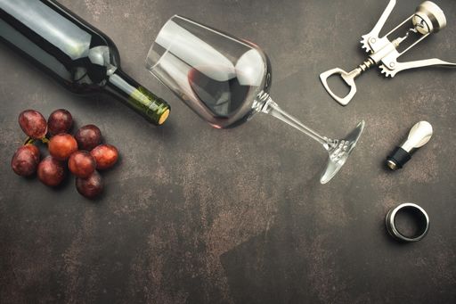A top view of a red wine glass and bottle on a stone background with a corkscrew and grapes