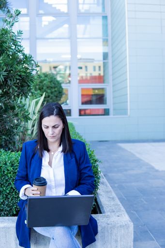 Beautiful Caucasian businesswoman using a laptop computer and holding a cup of coffee sitting outside an office building, copy space