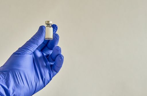 A doctor's hand in medical glove holding a small vial containing a vaccine