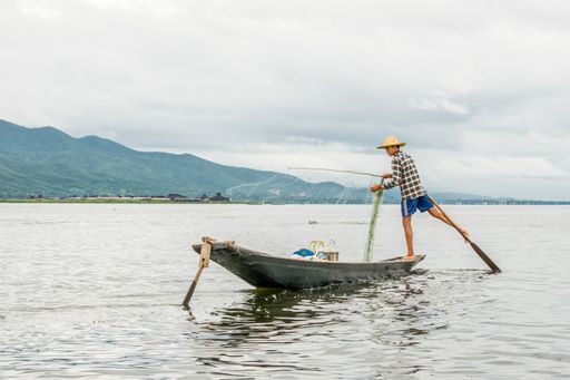 Local young Burmese male fisherman wearing checked shirt, using stick and net to fish, balancing on one foot on boat, Inle Lake Myanmar, Burma