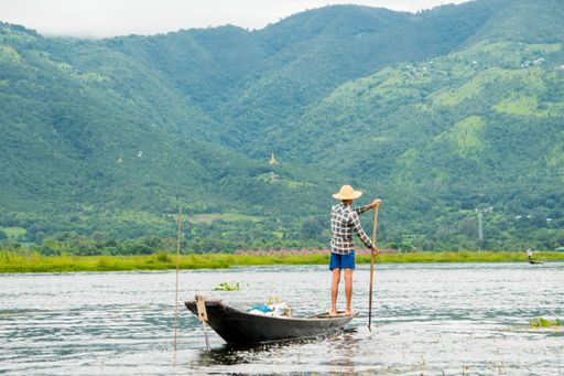 Local young Burmese fisherman wearing a checked shirt rowing after fishing, standing on the edge of a thin long boat, Inle Lake, Myanmar, Burma