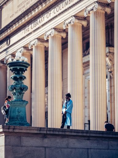 A vertical shot of a female taking a photo of her friend in a graduation gown in Columbia University