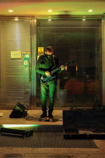 Busker playing a guitar in the evening in the alcove of a shop. Wearing a mask because of Covid-19 . Guitar case open for donations