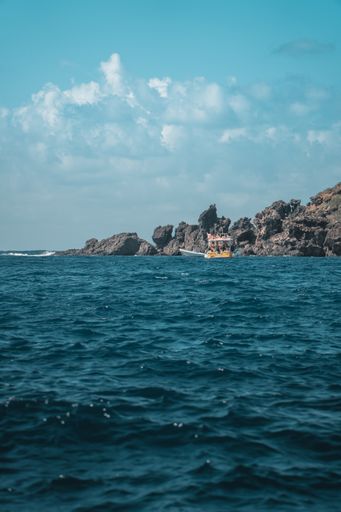 A vertical shot of a body of water near a rocky shore with  a small boat floating against a light blue sky