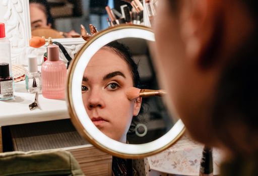 Selective focus Portrait of young woman in mirror doing her make-up. Woman using brush on her cheeks in front of circular led illuminated make-up mirr