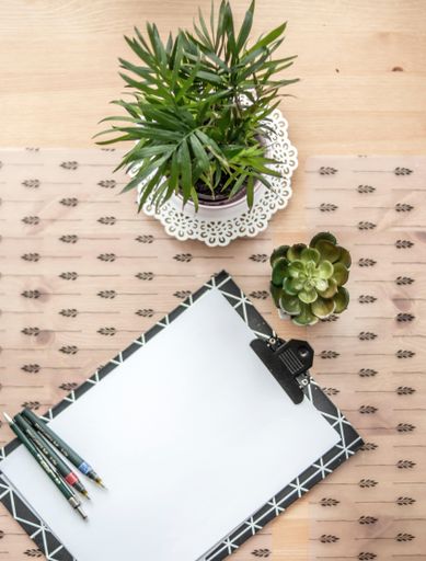 An overhead shot of a white paper with pens on a wooden surface with green plants