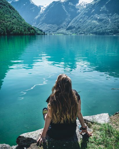 A vertical shot of a girl sitting on a stone near a mesmerizing blue lake among huge mountains