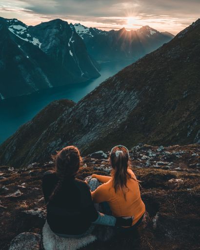 A vertical shot of two girls sitting on the slope of the mountain mesmerized by the view at sunset