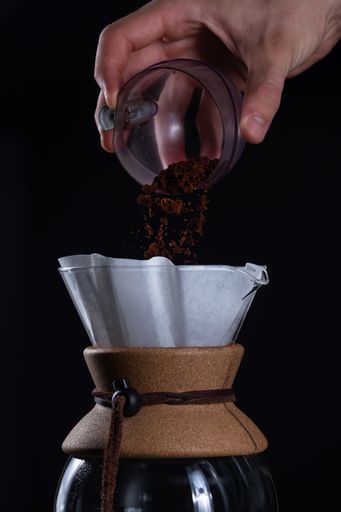 A vertical closeup of the hand adding fresh coffee grinds into a glass coffeemaker.