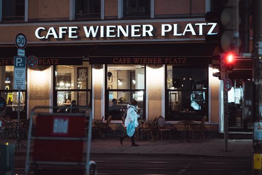 A photo of Cafe Wiener Platz in Munich at night, an old woman walks past the shop window