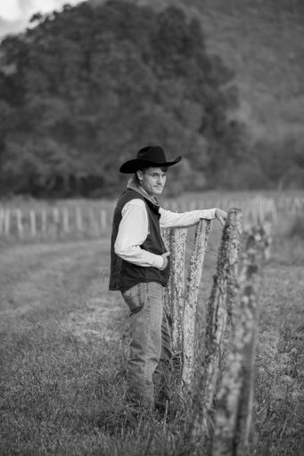 A vertical greyscale shot of a man wearing cowboy outfit and standing next to a fence in nature