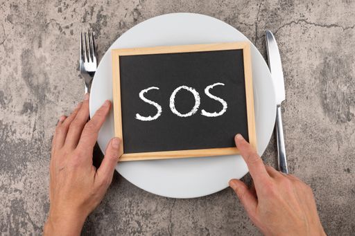 A photo of man's hands with fork, knife and an "SOS" word on a board on a plate