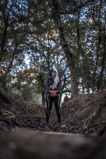A vertical shot of a female wearing a skeleton costume and skull makeup in a creepy forest