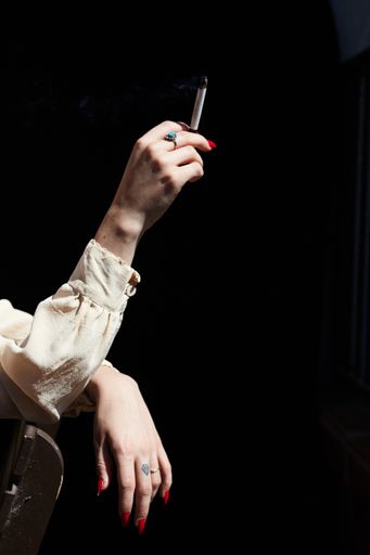 Feminine Woman's Hand Holding Marijuana THC CBD Joint with bright red nails on black background. Dramatic shadows in the sunlight.