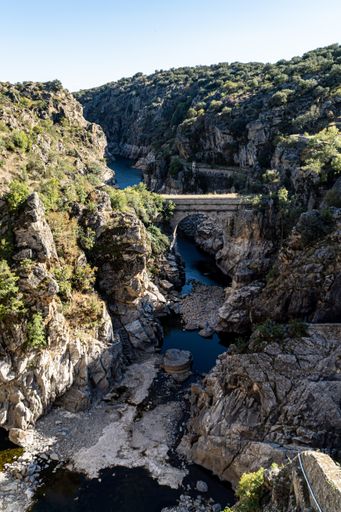 Vertical shot of a blue river flowing through a small valley or ravine, with an abandoned stone bridge and lots of green vegetation on the cliffs.
