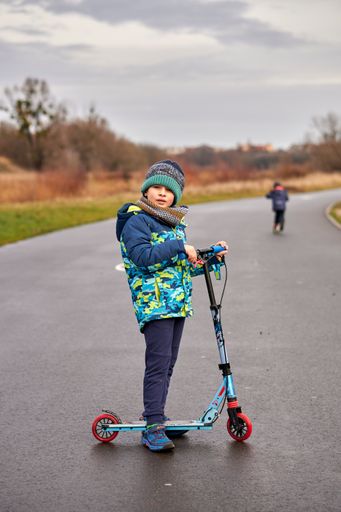 A Polish Caucasian boy standing with a scooter on a pathway next to the Warta river on a cloudy day.