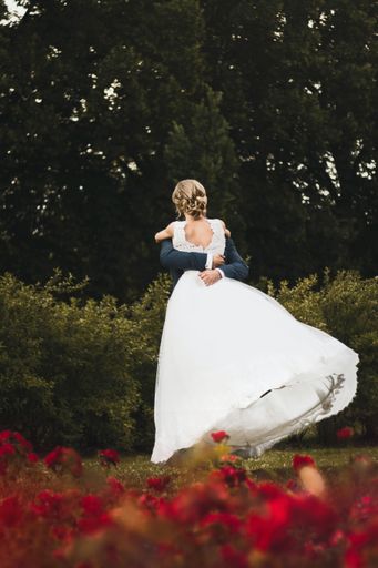 A vertical shot of newlywed couple hugging each other on red flowers foreground in a garden- perfect for newly married, newlywed, wedding concepts