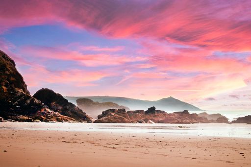 The beach surrounded by rocks and the sea under a cloudy sky during a beautiful pink sunset