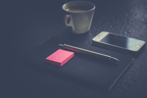 A selective shot of a gray pen and pink sticky notes on a black notebook near white cellphone and coffee mug