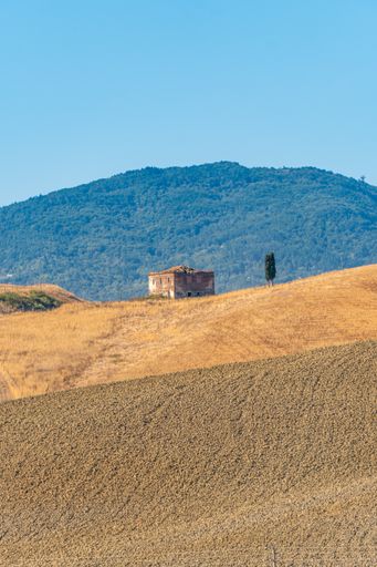 An old building in a steppe and forested mountains under a blue sky on a sunny day in Tuscany, Italy