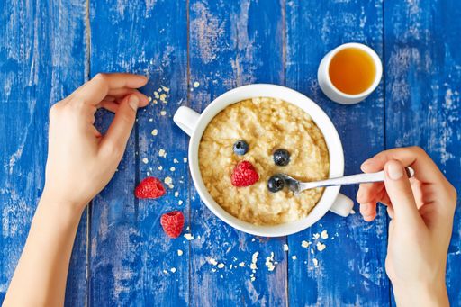 A top view of female hands eating oat porridge with berries from a white bowl and small white bowl of syrup on a wooden blue table