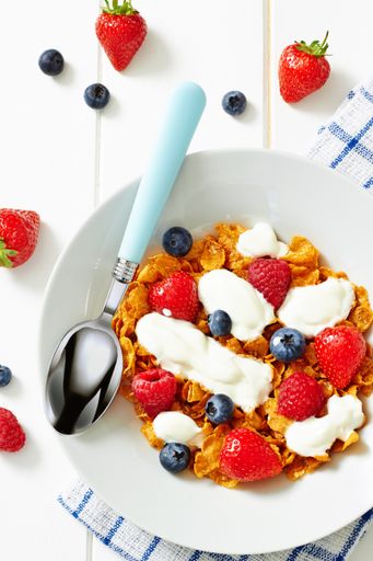 A top view of breakfast cereal with yogurt and fruits with a spoon in white bowl on wooden white table